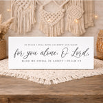 A white sign with black text that reads "In peace I will both lie down and sleep: for you alone, O Lord, Make me dwell in safety—PSALM 4:8" is displayed on a wooden surface.