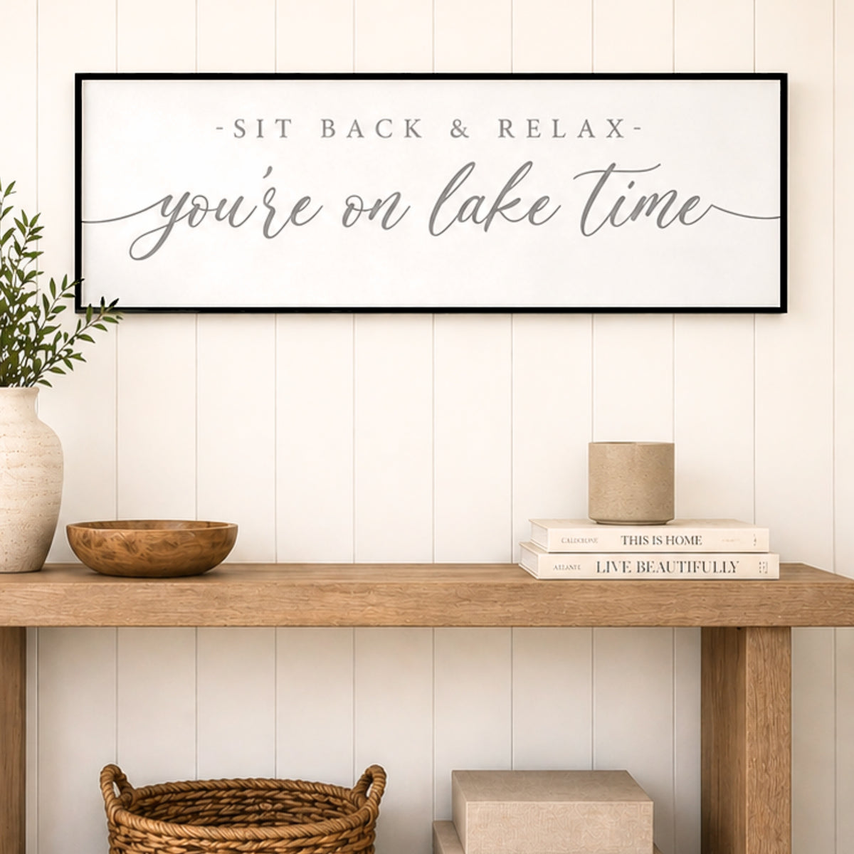 a wooden table with a framed sign that reads "SIT BACK & RELAX you're on lake time" above it. The table also has a small bowl, a stack of books, and a wicker basket.