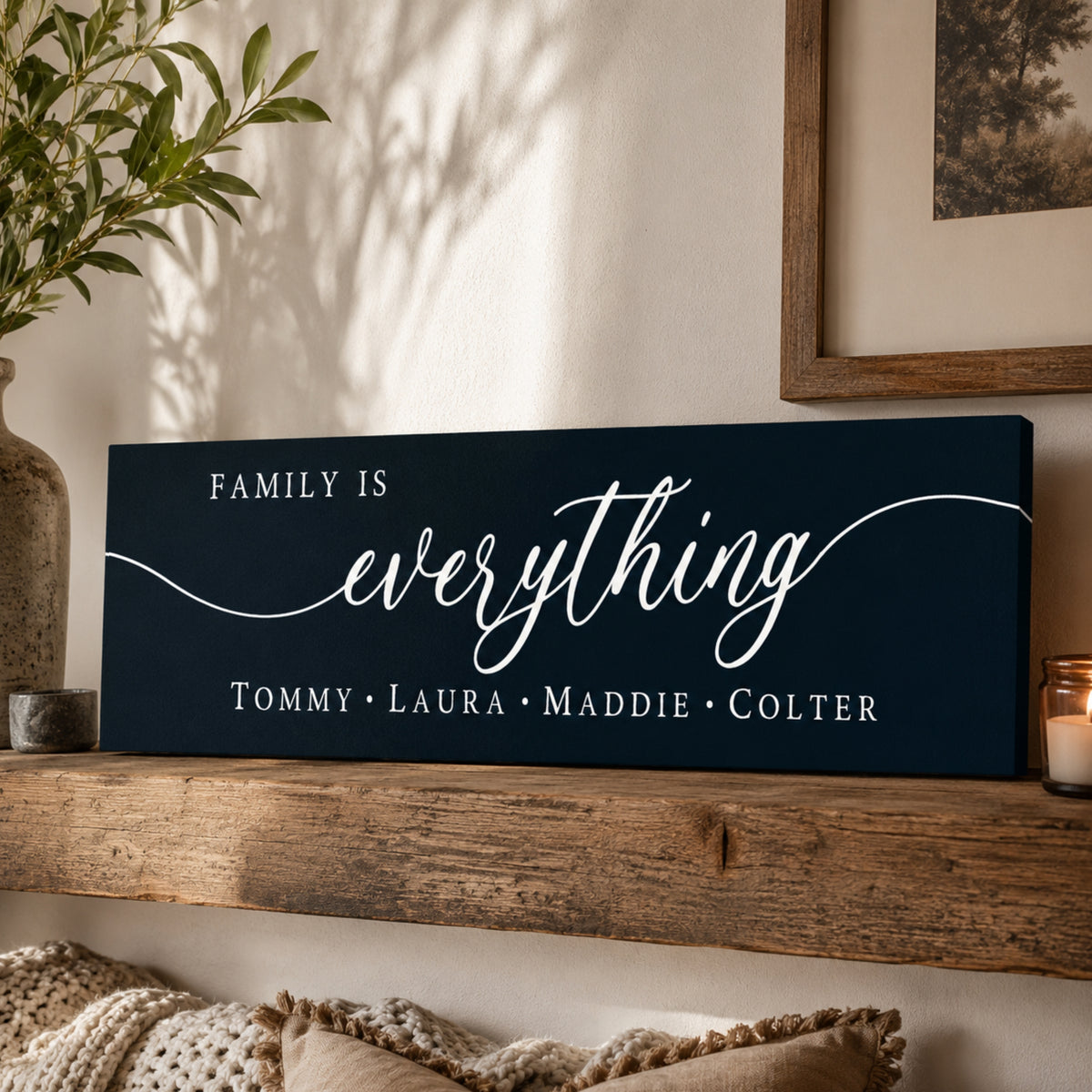 A dark blue sign with the text "Family is everything" in white cursive font, placed on a wooden shelf next to a plant and a candle.