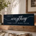 A dark blue sign with the text "Family is everything" in white cursive font, placed on a wooden shelf next to a plant and a candle.