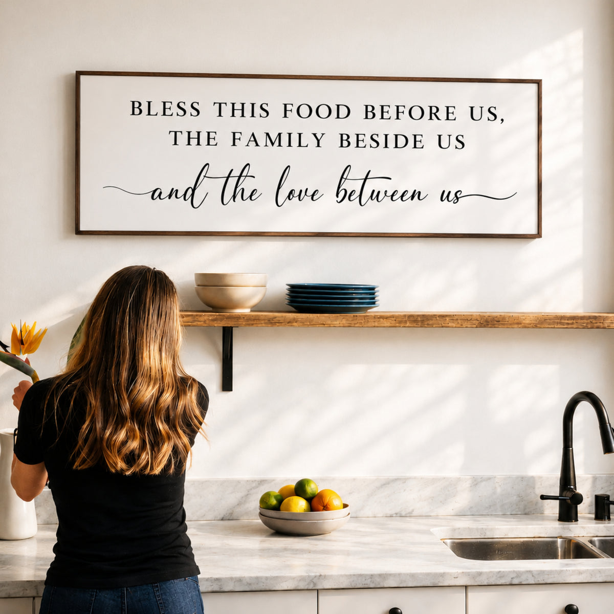 A woman stands in a kitchen, looking at a framed sign that reads "Bless this food before us, the family beside us and the love between us".