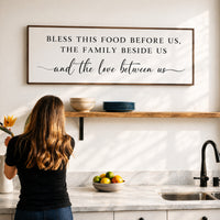 A woman stands in a kitchen, looking at a framed sign that reads "Bless this food before us, the family beside us and the love between us".
