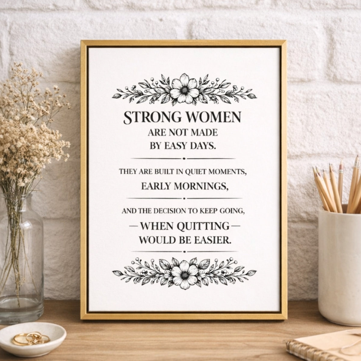 a framed inspirational quote with a floral border, placed on a wooden surface next to a vase with flowers and a cup with pencils.