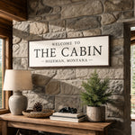 a wooden table with a lamp, a potted plant, and a framed sign that reads "Welcome to The Cabin Bozeman, Montana".
