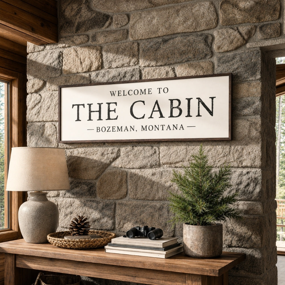 a wooden table with a lamp, a potted plant, and a framed sign that reads "Welcome to The Cabin Bozeman, Montana".