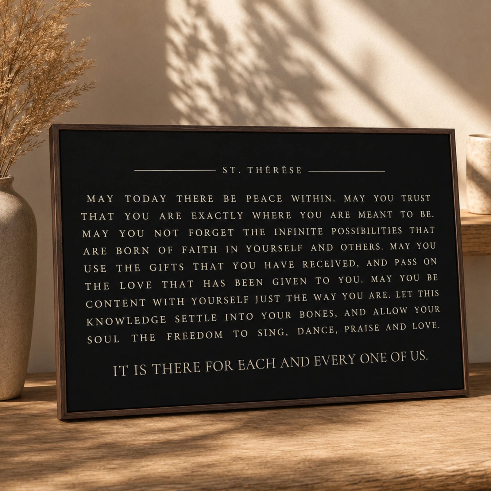 a framed quote on a wooden surface, with a vase of dried flowers to the left.