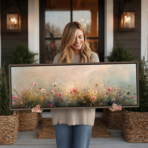 A woman is holding a large framed painting of a field of flowers in front of a house.