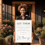 A woman is standing in front of a wooden door, holding a framed sign with the message "Let Them Go" written on it.