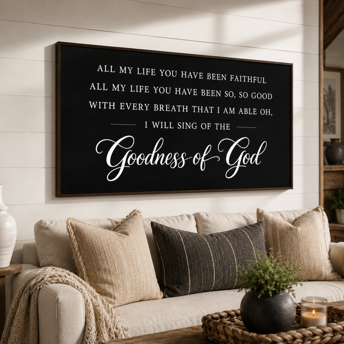 a black framed sign with white text that reads "All my life you have been faithful, all my life you have been so, so good with every breath that I am able oh, I will sing of the Goodness of God" displayed on a wall above a couch with pillows and a potted plant.