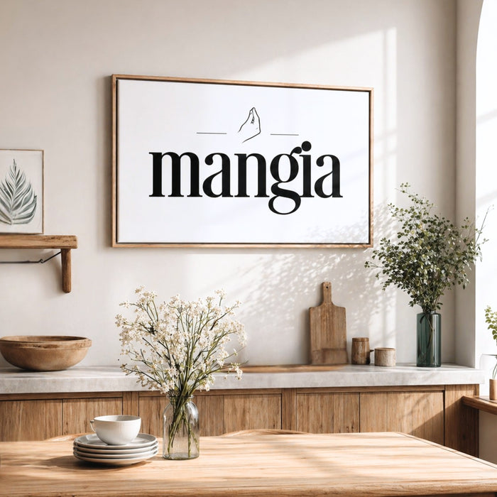 a kitchen counter with a framed sign that reads "mangia" above it. On the counter, there are a vase of flowers, a bowl, and a wooden cutting board. The overall scene has a warm and inviting atmosphere.