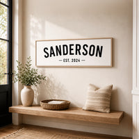 a wooden bench with a basket and a plant on it, set against a wall with a framed sign that reads "SANDERSON EST. 2024".