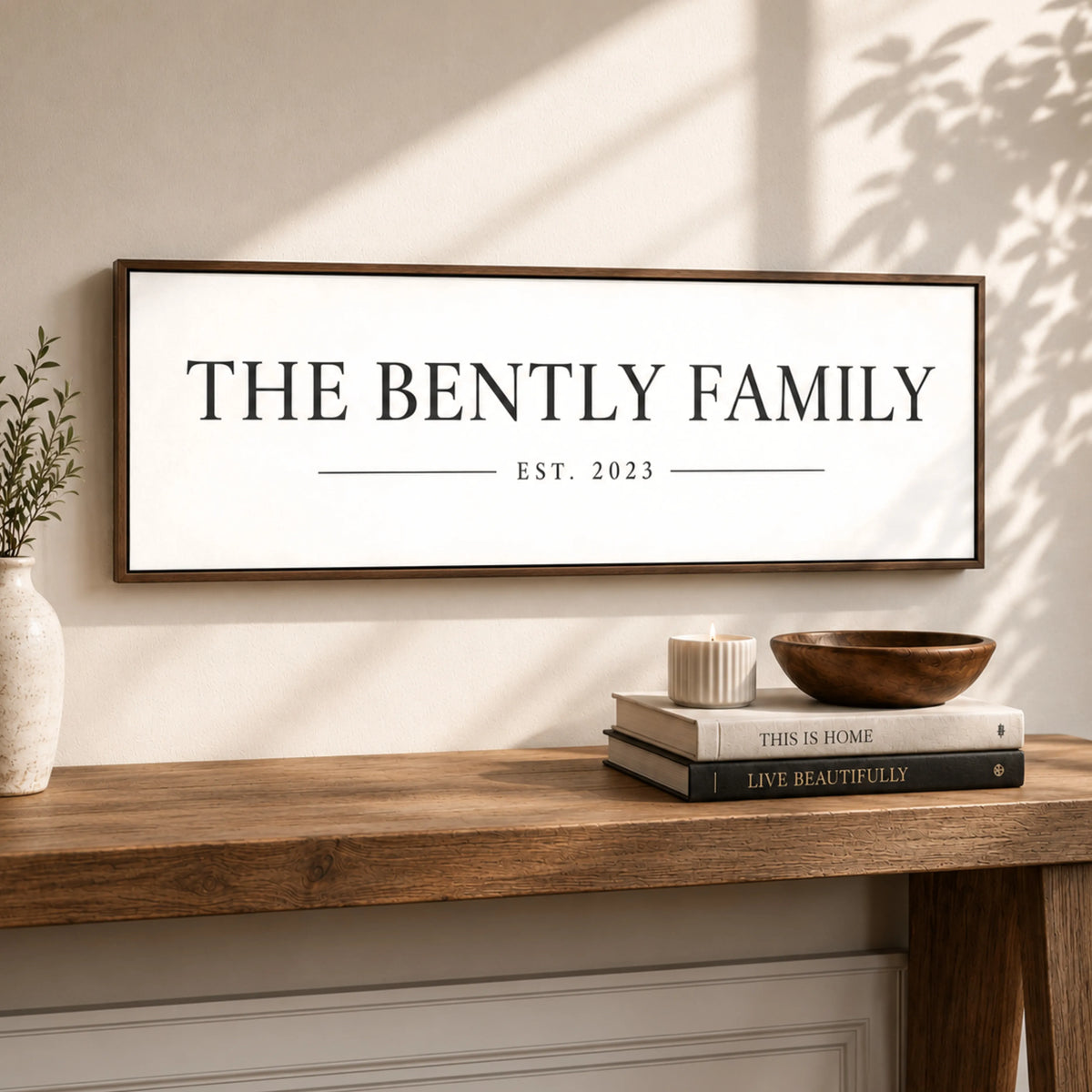 a wooden table with a framed sign that reads "THE BENTLY FAMILY EST. 2023" above it. There are also two books and a bowl on the table.