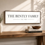 a wooden table with a framed sign that reads "THE BENTLY FAMILY EST. 2023" above it. There are also two books and a bowl on the table.