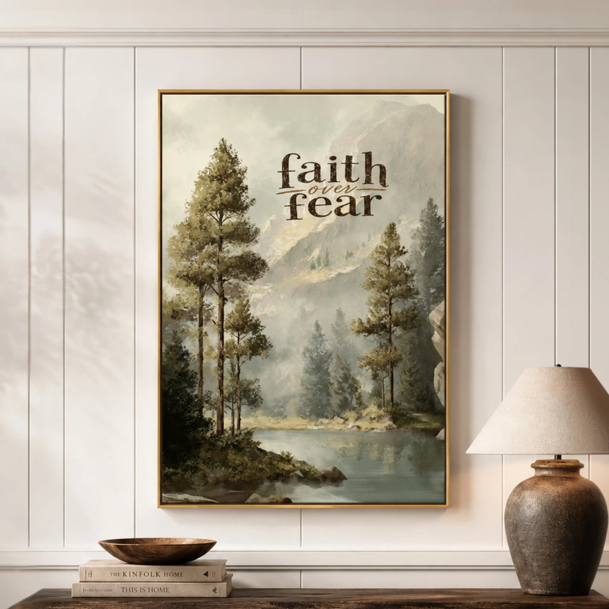 a framed painting of a serene landscape with trees, a lake, and mountains in the background, hanging on a wall above a wooden table with a lamp and a stack of books.