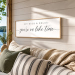 A framed sign on a wall that reads "SIT BACK & RELAX you're on lake time" with a scenic view of a lake and trees in the background.