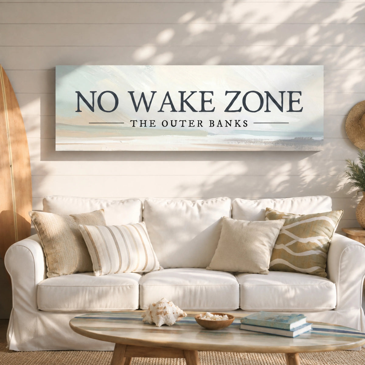 a living room with a white sofa, a coffee table, and a surfboard on the wall. Above the sofa, there is a large sign that says "NO WAKE ZONE THE OUTER BANKS".