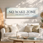a living room with a white sofa, a coffee table, and a surfboard on the wall. Above the sofa, there is a large sign that says "NO WAKE ZONE THE OUTER BANKS".