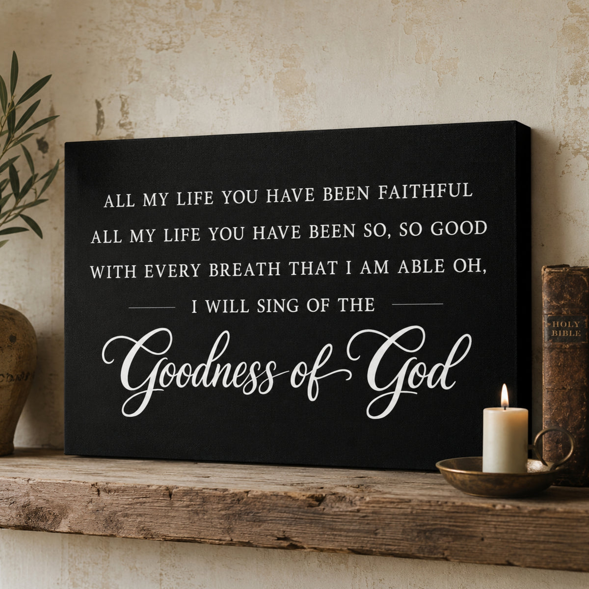 a black canvas with a white inscription that reads "All my life you have been faithful all my life you have been so, so good with every breath that I am able oh, I will sing of the Goodness of God". The canvas is displayed on a wooden shelf, with a lit candle and a small book to the right of it.