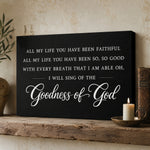 a black canvas with a white inscription that reads "All my life you have been faithful all my life you have been so, so good with every breath that I am able oh, I will sing of the Goodness of God". The canvas is displayed on a wooden shelf, with a lit candle and a small book to the right of it.