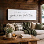 a cozy living room with a wooden wall, a large white sign with a message, a couch with pillows and a green blanket, a wicker basket on a coffee table, and a candle on a tray.
