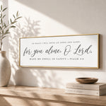 A framed Bible verse, "In peace I will both lie down and sleep; for you alone, O Lord, Make me dwell in safety—PSALM 4:8", is displayed on a wall above a wooden shelf with a stack of books and a plant.